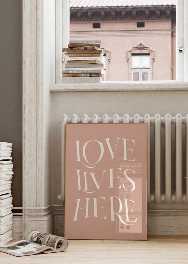 decoración con cuadros, ideas - Cuadro con frase "Love Lives Here" con fondo rosado, estilo nórdico. Una obra para colgar en cualquier estancia de la casa.